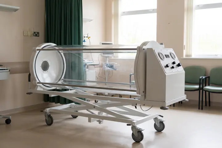 A clear acrylic monoplace hyperbaric chamber on a wheeled base in a hospital treatment room